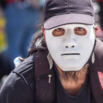 Fotografía de un hombre vestido de negro, con un morral en los hombros, que se ve que está agachado y que tiene una careta blanca que le tapa el rostro.