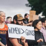 Hombre con gorra sostiene un letrero con el texto "WOMEN > GUNS" entre una multitud en una manifestación al aire libre. A su lado, una mujer con camiseta naranja de Denver. Al fondo, más personas y árboles.