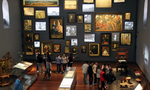 Foto panorámica de la Sala Memoria y nación del Museo Nacional de Colombia en la que se ven varios objetos colagos de la pared y muchas personas visitando el sitio.