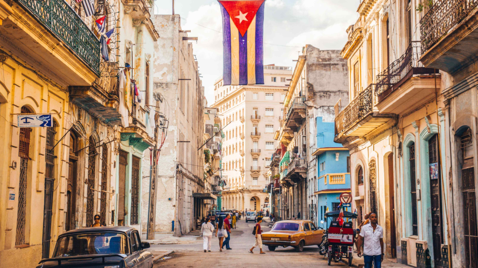 Callejón de La Habana, colorido, con carros viejos y la bandera cubana.