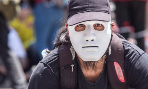 Fotografía de un hombre vestido de negro, con un morral en los hombros, que se ve que está agachado y que tiene una careta blanca que le tapa el rostro.