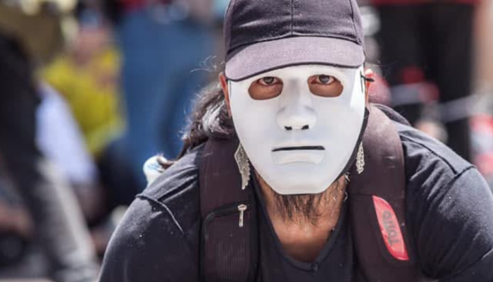 Fotografía de un hombre vestido de negro, con un morral en los hombros, que se ve que está agachado y que tiene una careta blanca que le tapa el rostro.
