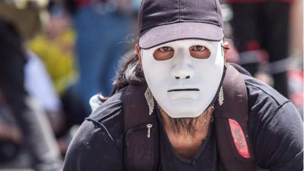Fotografía de un hombre vestido de negro, con un morral en los hombros, que se ve que está agachado y que tiene una careta blanca que le tapa el rostro.