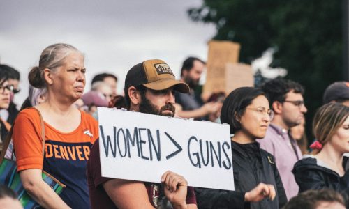Hombre con gorra sostiene un letrero con el texto "WOMEN > GUNS" entre una multitud en una manifestación al aire libre. A su lado, una mujer con camiseta naranja de Denver. Al fondo, más personas y árboles.