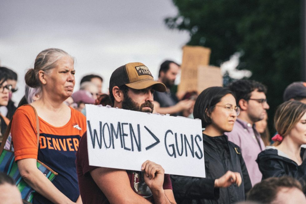 Hombre con gorra sostiene un letrero con el texto "WOMEN > GUNS" entre una multitud en una manifestación al aire libre. A su lado, una mujer con camiseta naranja de Denver. Al fondo, más personas y árboles.